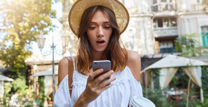 woman wears stylish hat and white summer dress, feels surprised, using smartphone