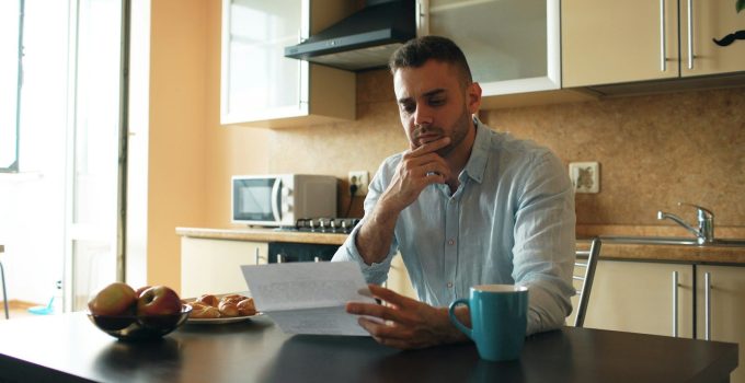 Man in a blue shirt sitting at a kitchen table, hand on chin, reading a paper document beside a blue mug