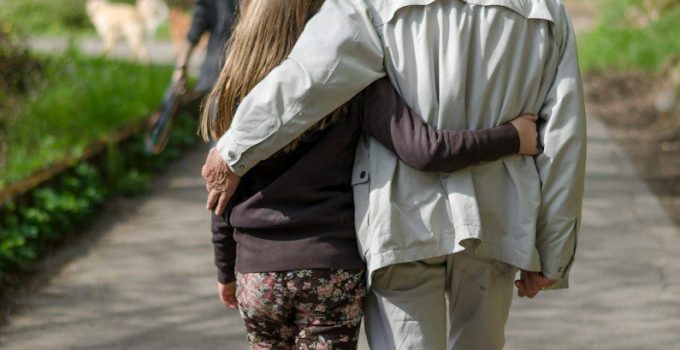 An older man and his adult daughter walking arm in arm along a tree-lined path, seen from behind