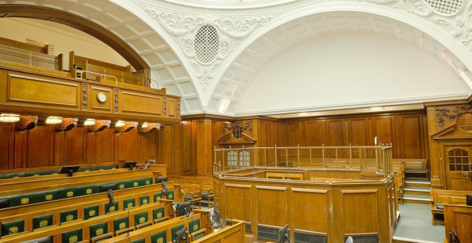 Empty ornate courtroom interior with wooden paneling, rows of wooden benches and green seats under a vaulted ceiling